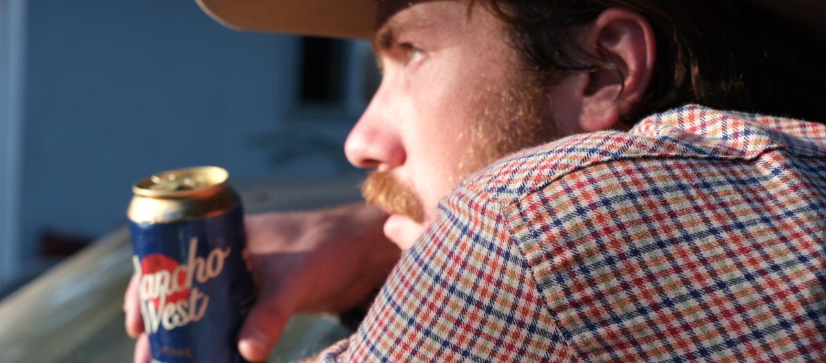 Man in a red, navy and tan check western shirt and straw hat, holding a Rancho West beer can, in late golden light